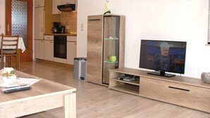 A modern living room with a wooden entertainment unit, a flat-screen TV, and a kitchen area in the background. - Haus Gertrud (Wittmund)