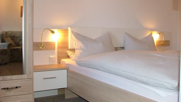 A bedroom with a bed, nightstands, a drawer, and wall-mounted lamps. - Haus Gertrud (Wittmund)