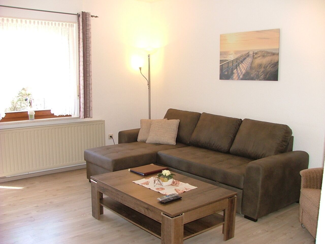 A living room with a brown sofa, a wooden coffee table with a remote control, a lamp, and a painting on the wall.