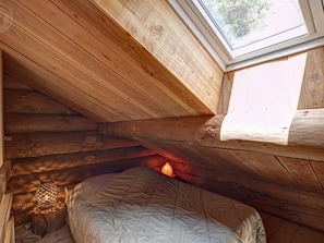 Interior de una cabaña de madera con techo inclinado, tragaluz, cama y lámpara de estilo farol.