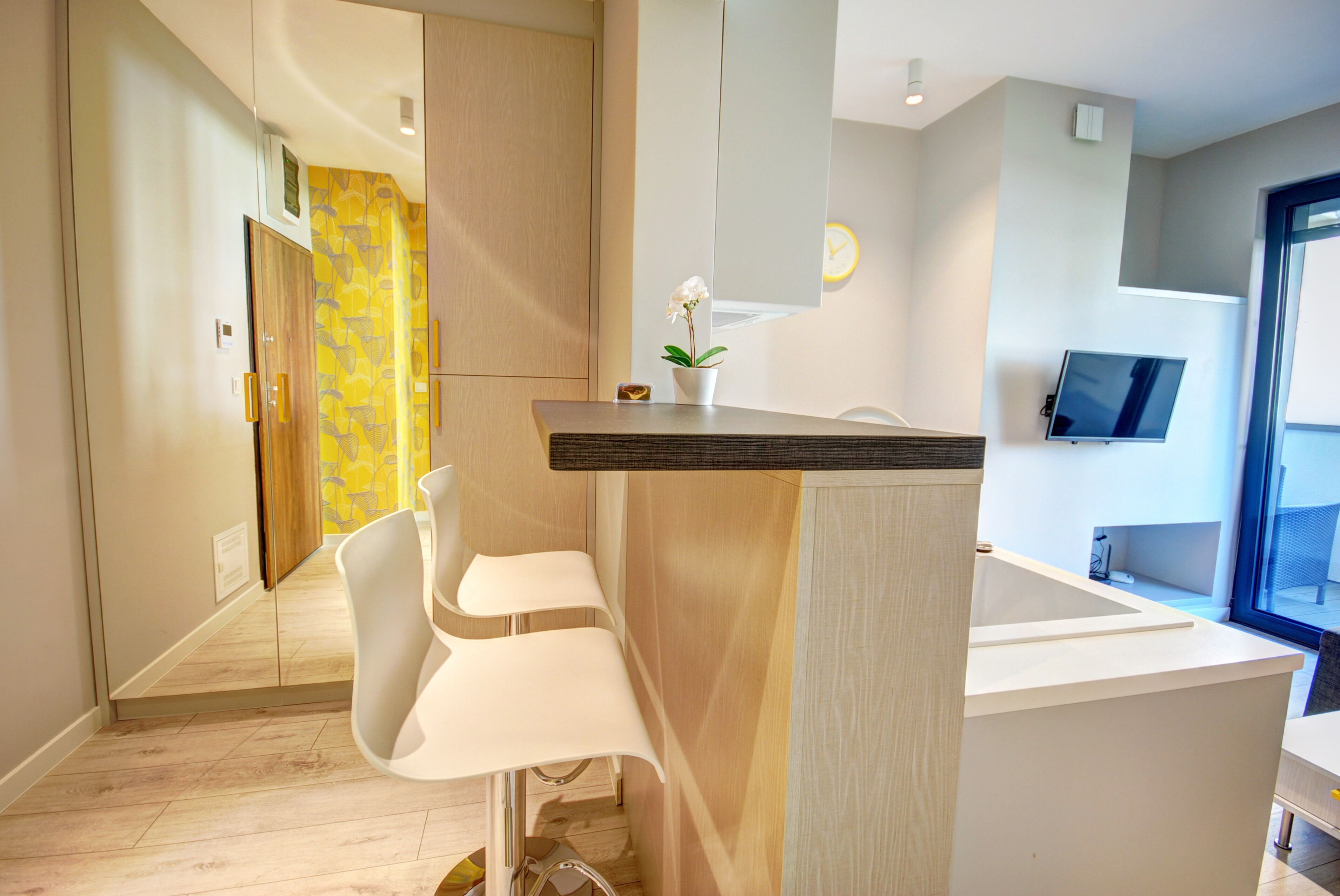 A modern hotel room with a wooden floor, a white desk with a plant, a white chair, and a TV on the wall.