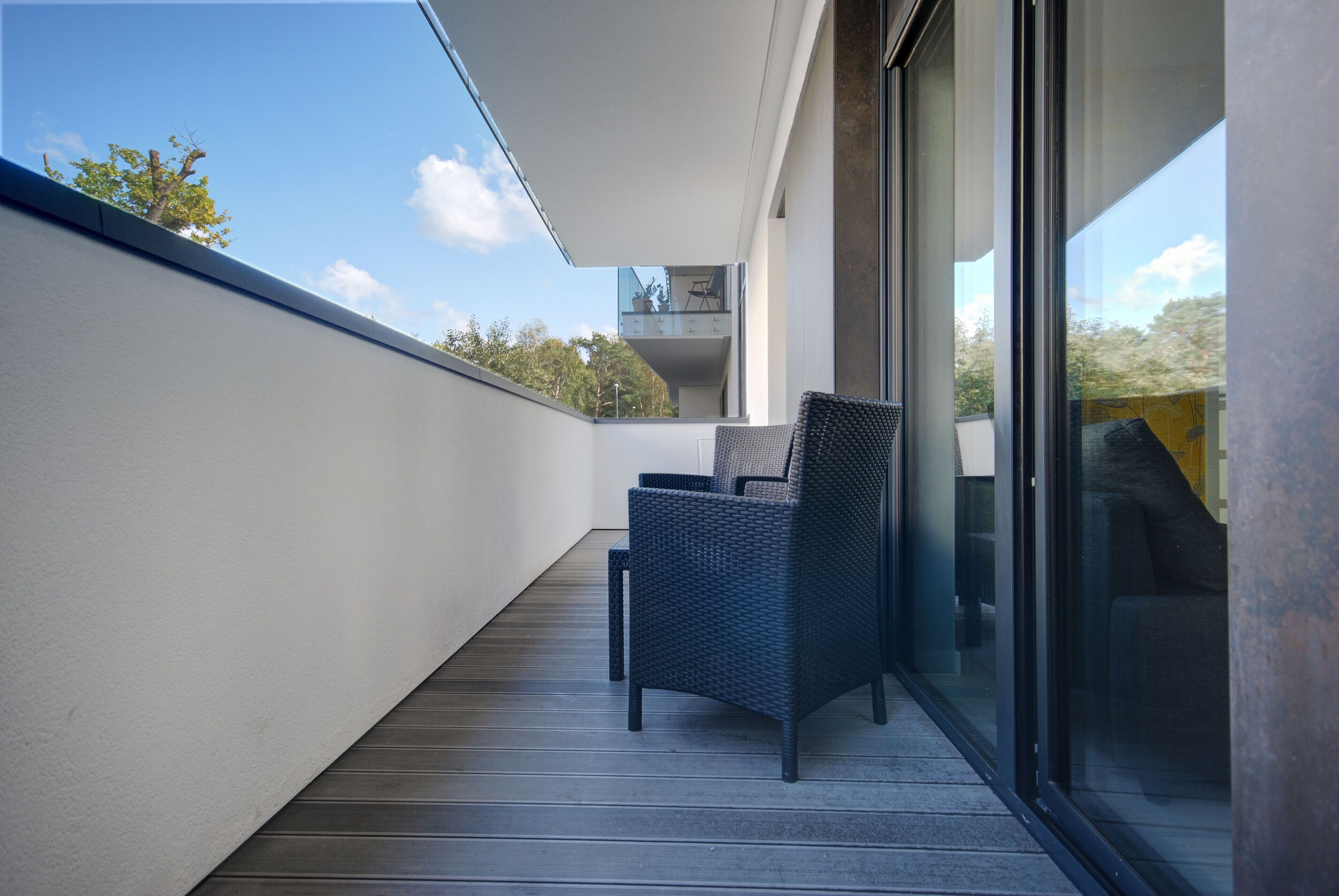 A balcony with a wicker chair and a small table, overlooking a modern building and trees.