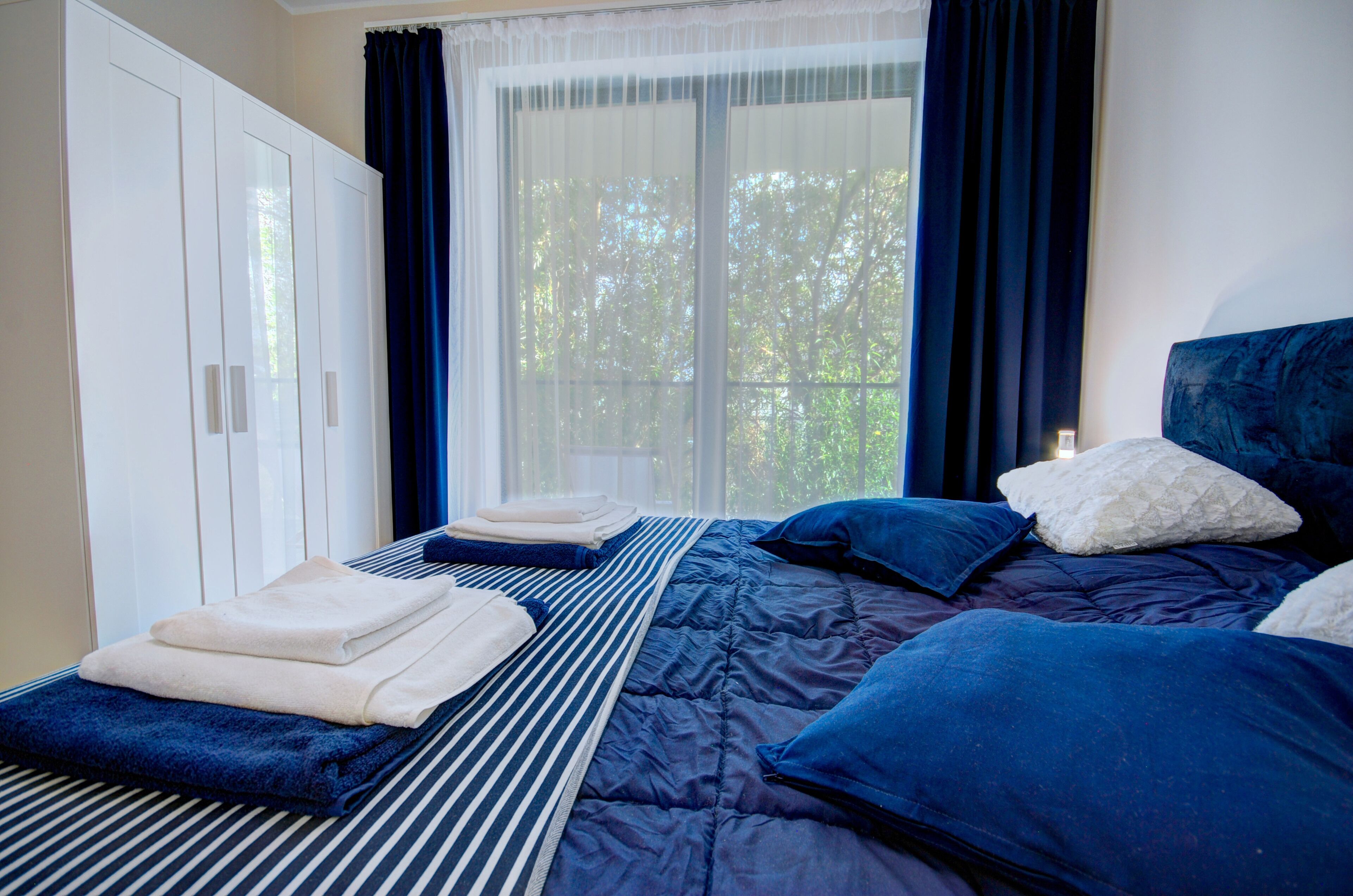 A bedroom with a large window, a bed with blue bedding, and a wardrobe.