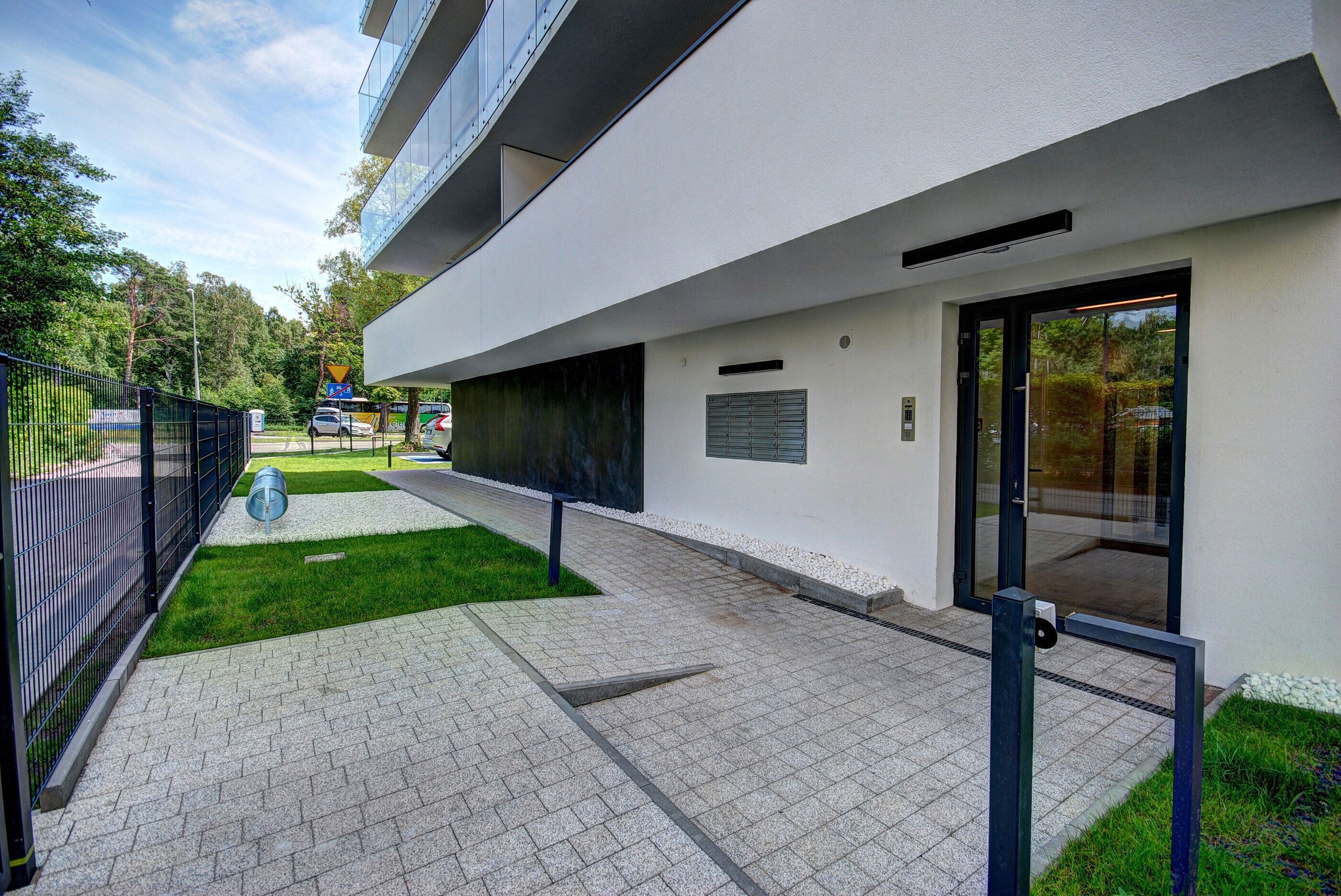Un edificio moderno con un sendero pavimentado, una cerca metálica y una zona ajardinada.