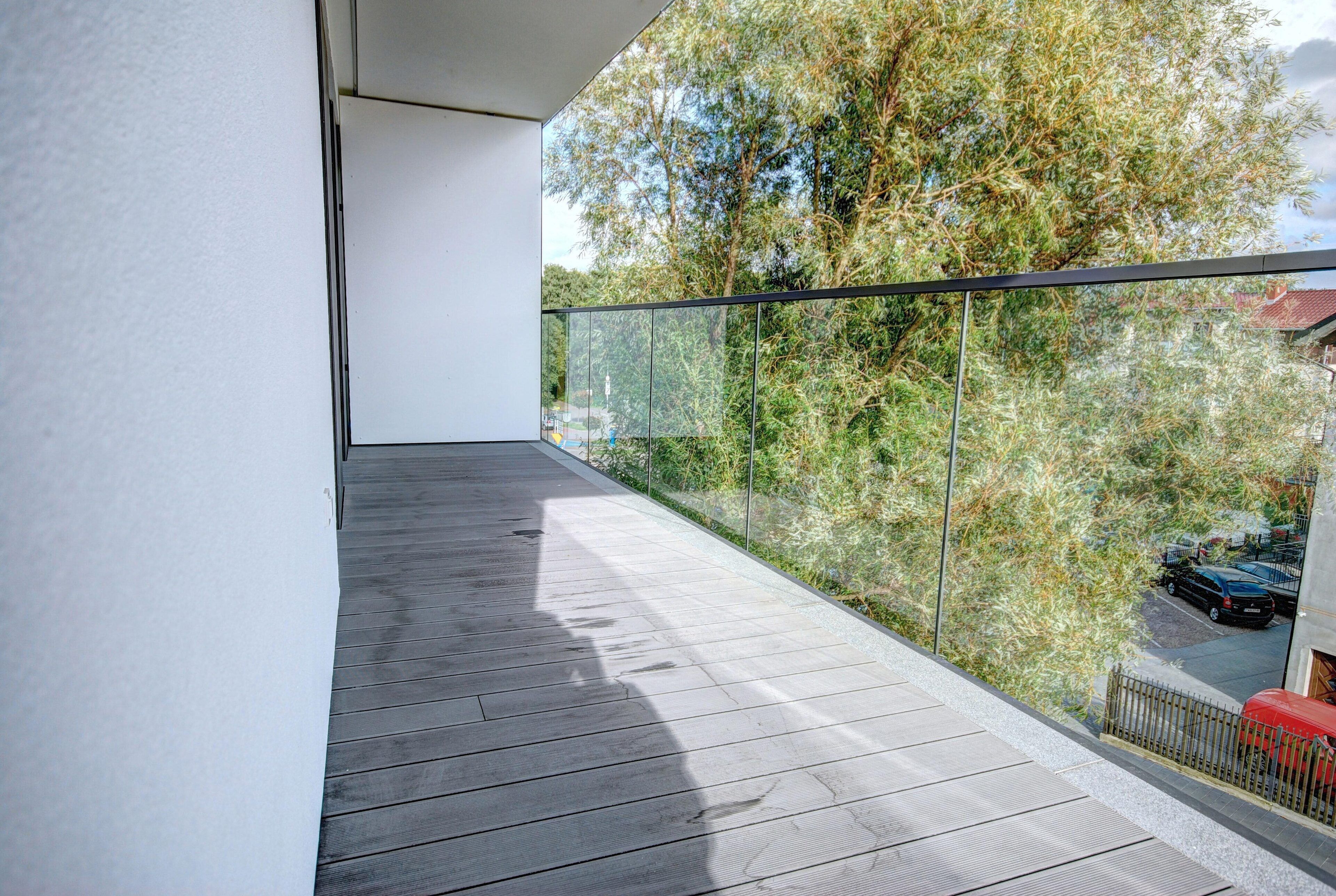 A balcony with a glass railing, a wooden floor, and a view of trees and a parked car.