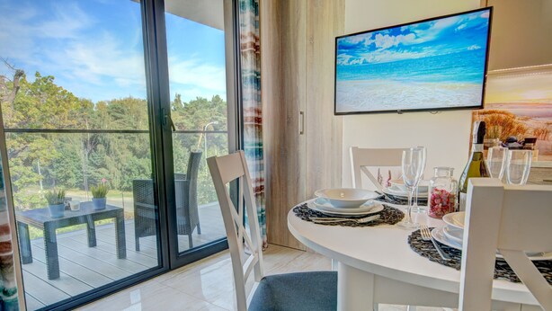 A dining area with a table set for two, a TV mounted on the wall, and a balcony with a view of greenery and the sky. - HelApart - NATURALnie że Hel (Hel)