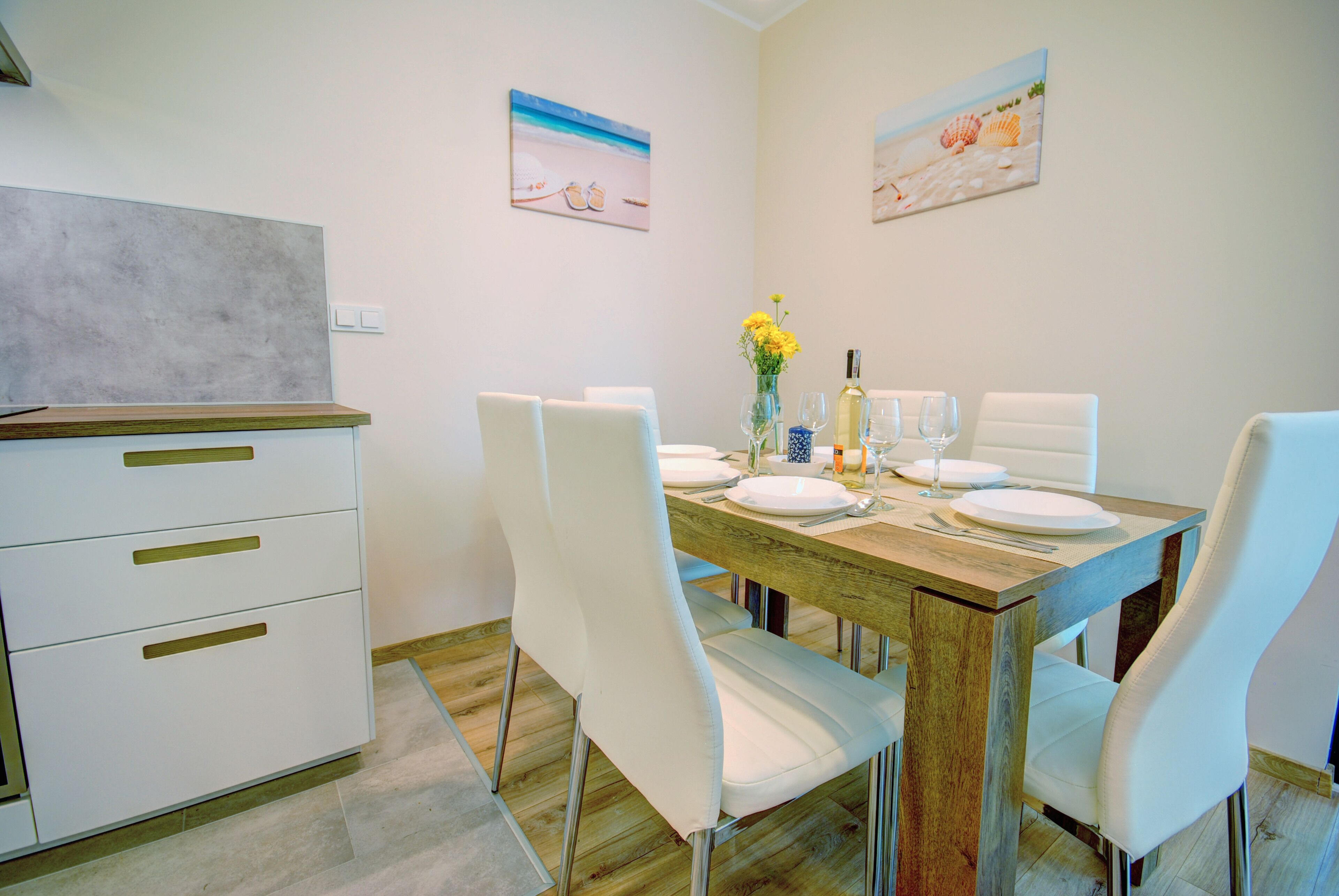 Un comedor con una mesa de madera dispuesta para cuatro, sillas blancas y una encimera de cocina con cajones.