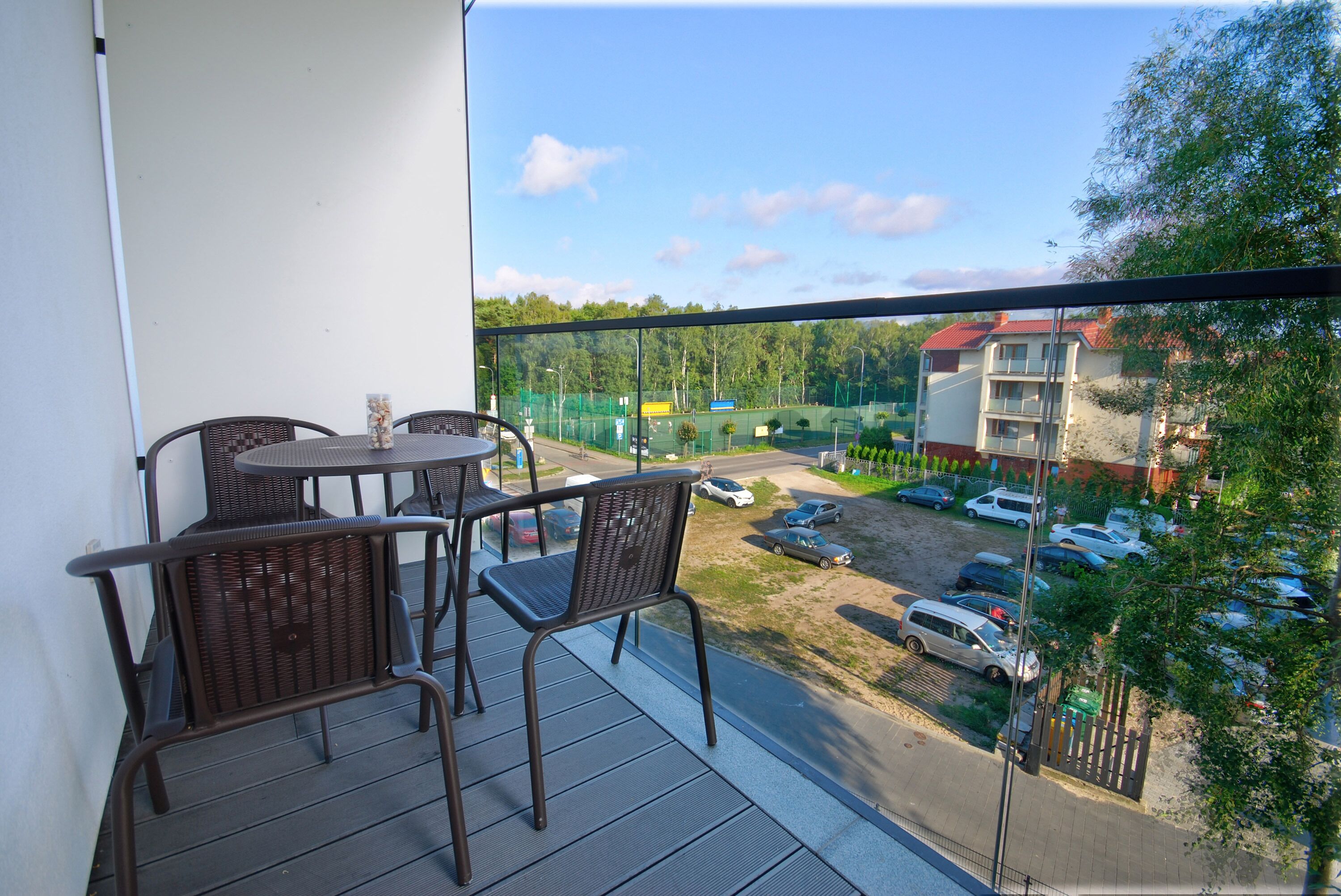 A balcony with wicker chairs and a table, overlooking a parking area and buildings.