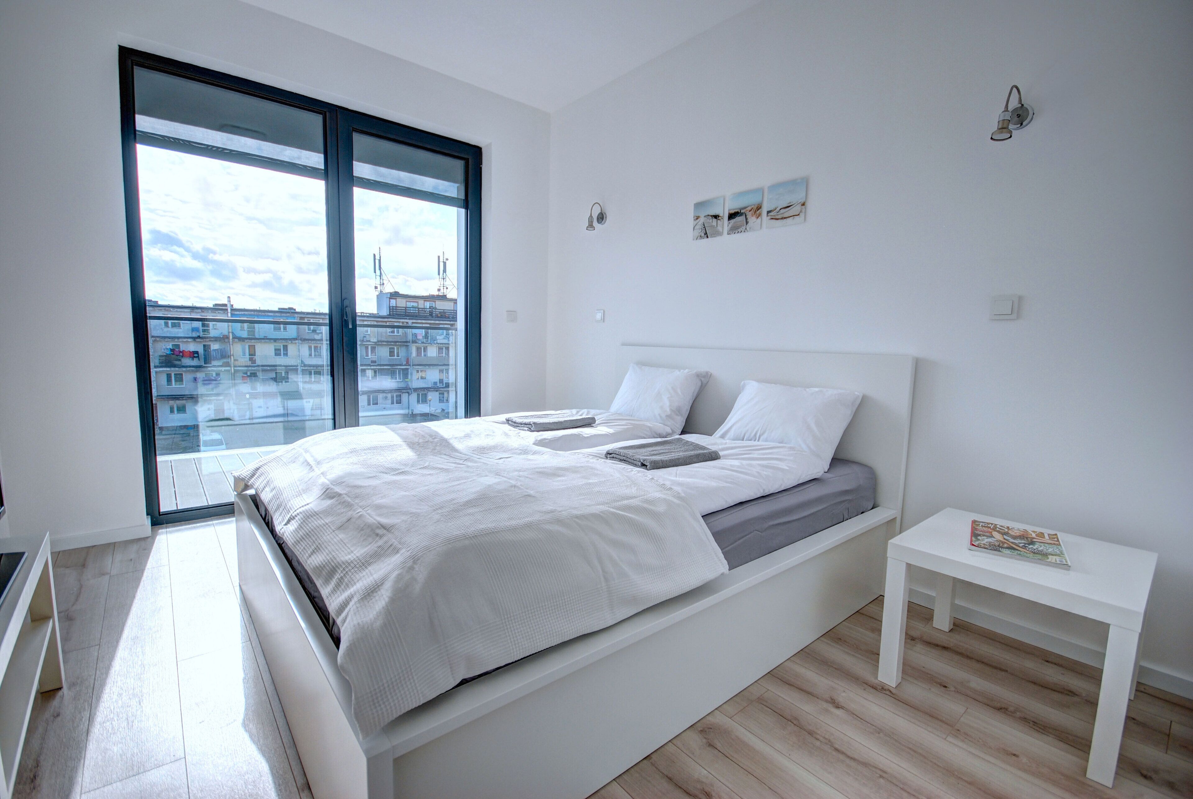 A modern bedroom with a large bed, a small bedside table, and a view of buildings through a large window.