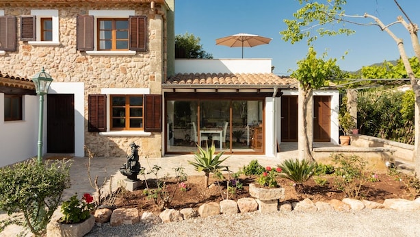 A stone house with a tiled roof, a garden with plants and a statue, and a clear blue sky.