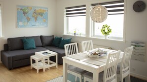 A modern living room with a grey sofa, white dining table, and chairs. A world map is on the wall, and there is a window with blinds and a hanging light fixture. - Mørk Apartments (Tórshavn)