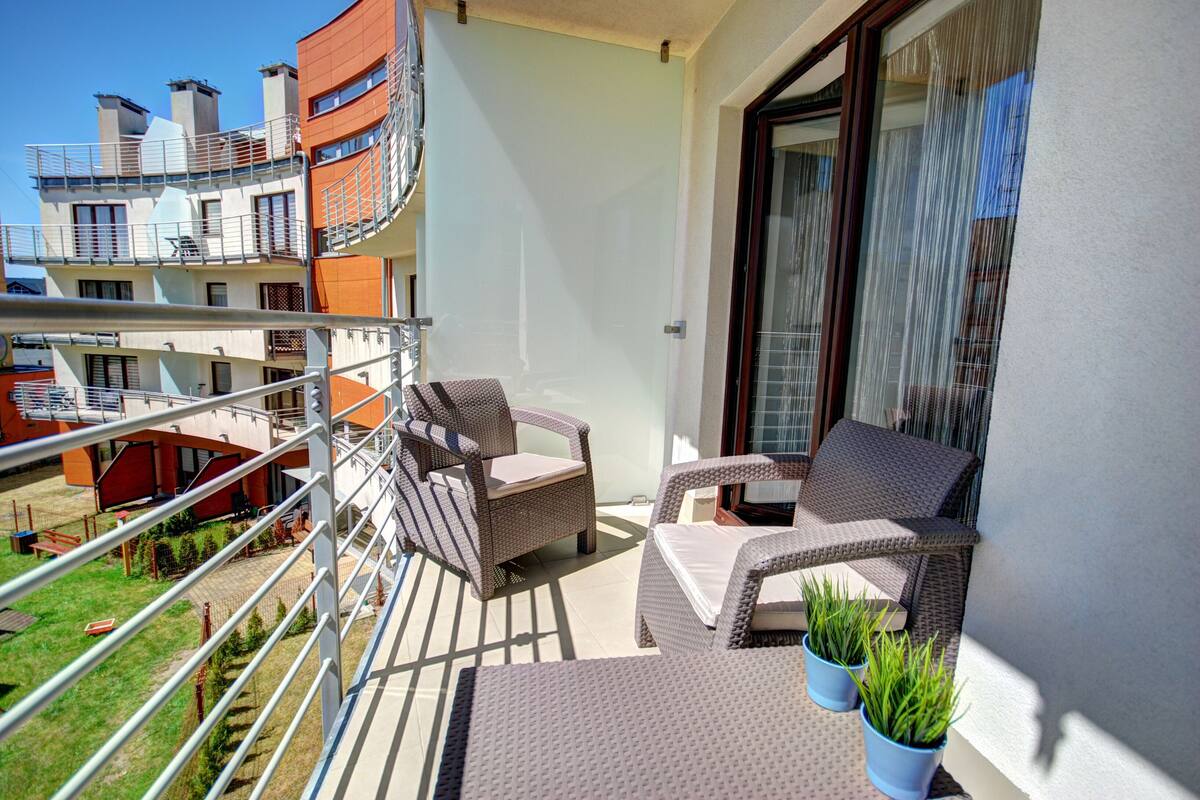 A balcony with wicker furniture and potted plants, overlooking a residential area with modern buildings.