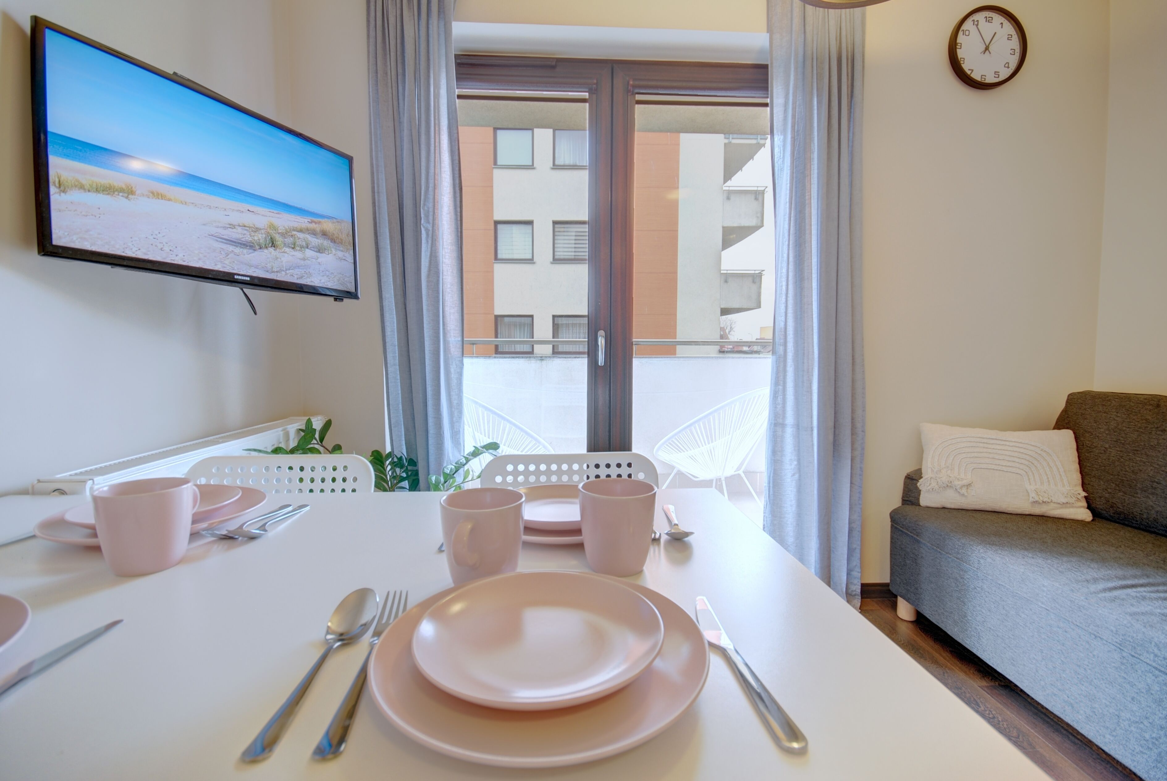 Et pænt anrettet spisebord med lyserøde tallerkener, bestik og kopper, et fladskærms-tv på væggen, en sofa og en balkon med udsigt til bygninger.