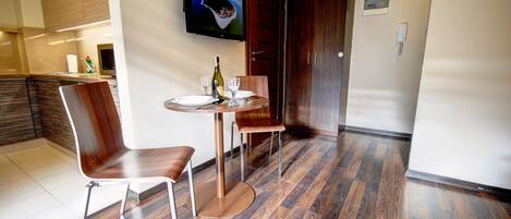 A dining area with a round table, two chairs, a bottle, and glasses. There is a TV mounted on the wall above the table.