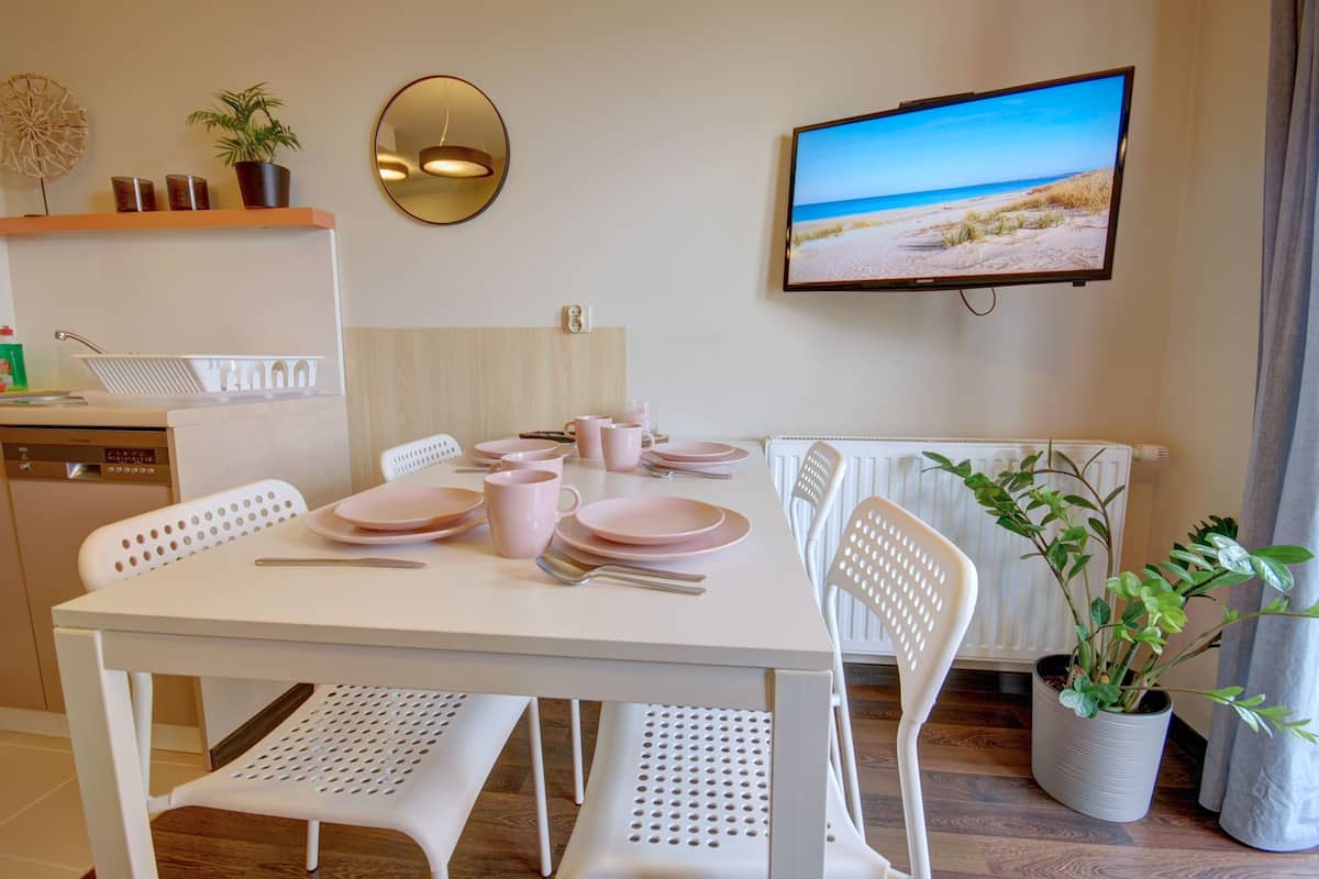 A dining area with a white table set for four, surrounded by white chairs. A television mounted on the wall displays a beach scene.