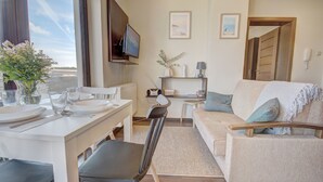 A modern living room with a dining area, a sofa, a TV mounted on the wall, and a view of the outdoors. - HelApart - Apartamenty Foka (Hel)