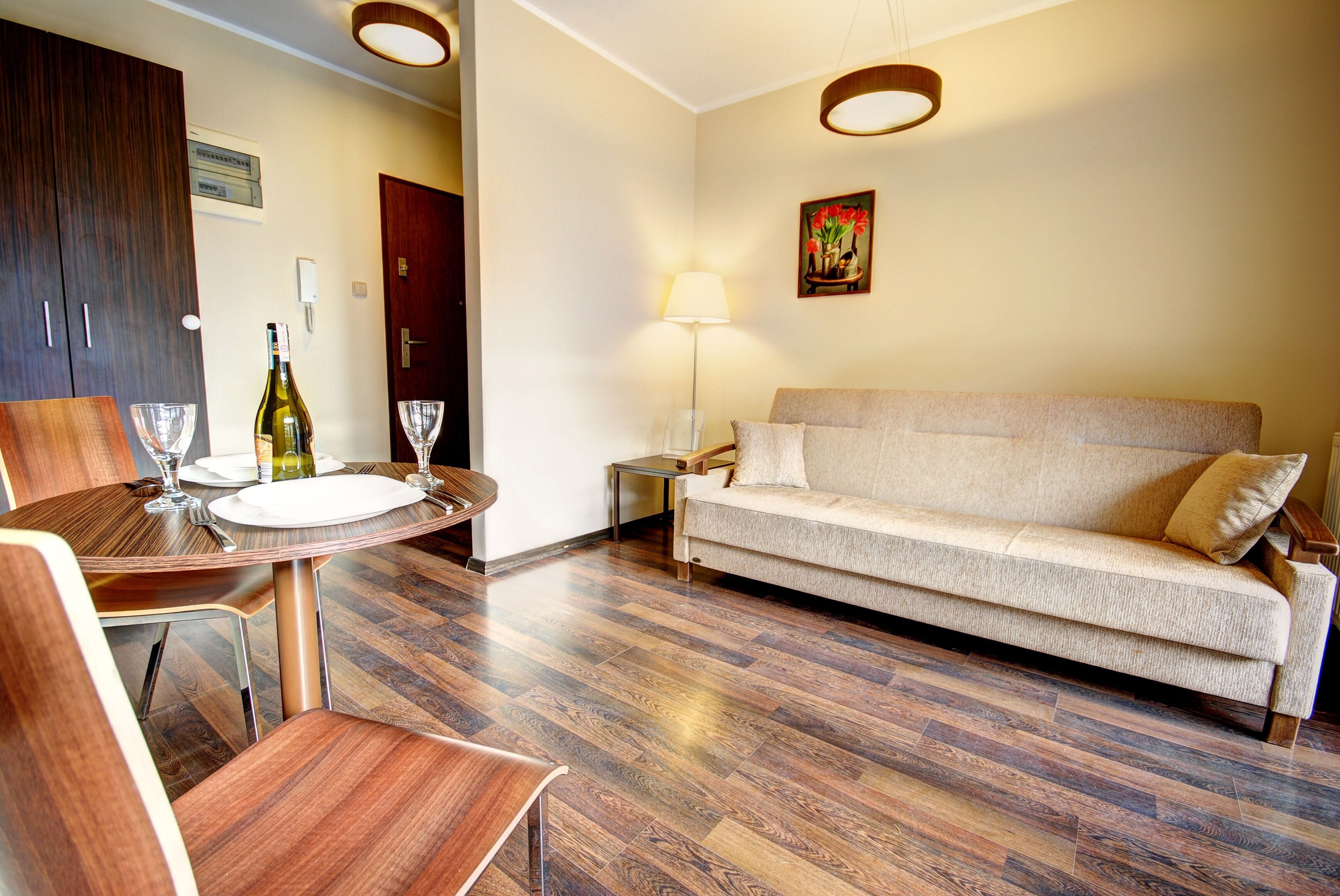 A dining area with a round table, chairs, and a bottle of wine. There is a sofa and a lamp in the living area.