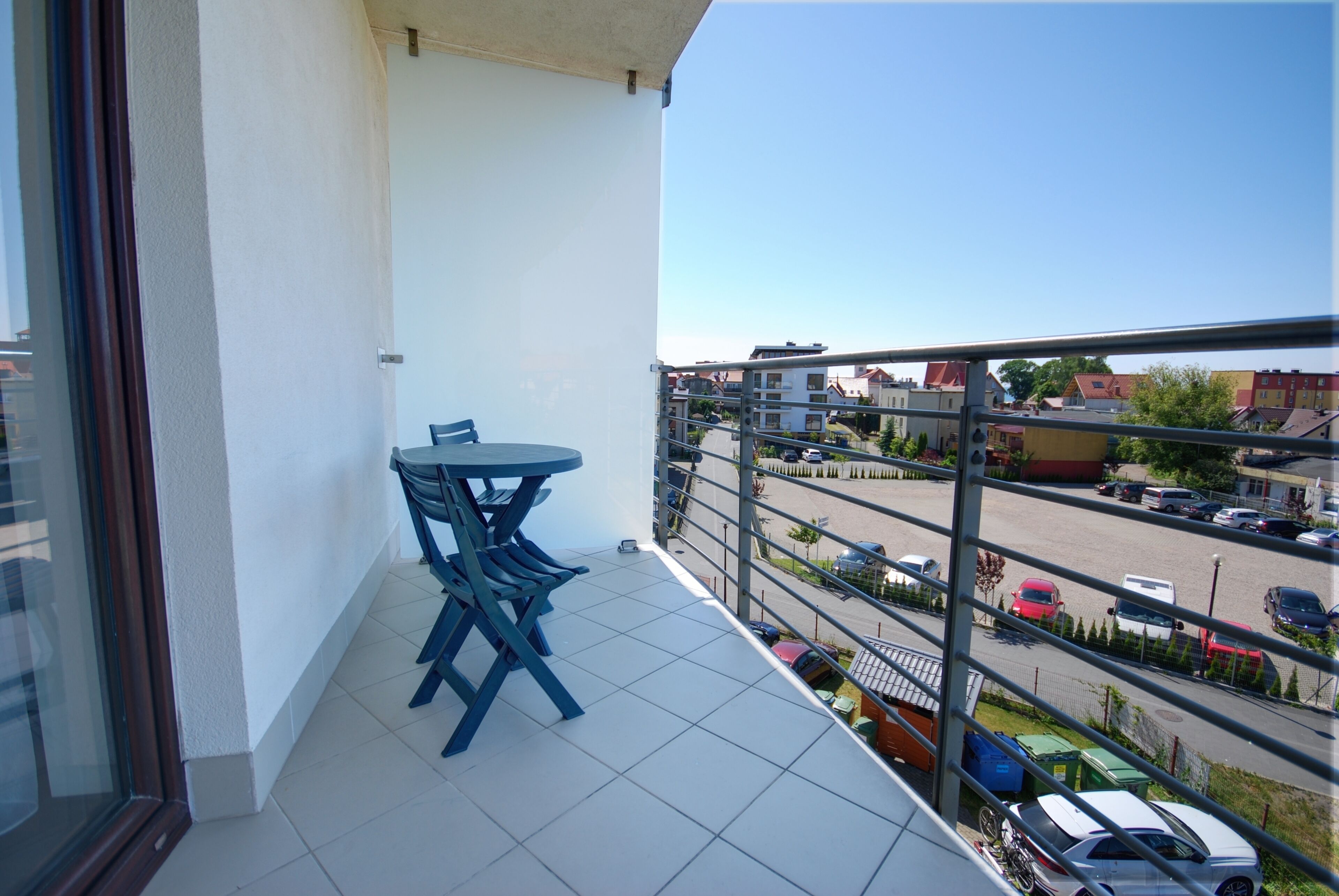 A balcony with a table and chairs, overlooking a parking area and buildings.