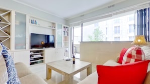 Ein modernes Wohnzimmer mit einem Flachbildfernseher, einem Holztisch und einer roten Sofaausstattung. Ein Bücherregal beherbergt Bücher und Dekoration, und ein großes Fenster bietet Blick auf die Gebäude draußen.