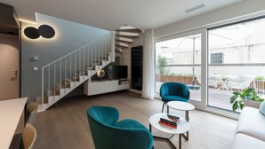 A modern living room with a staircase, teal chairs, a small white table, and a large window offering a view of the outdoors. - The Central City - Luxury ApartHotel (Luxembourg City)