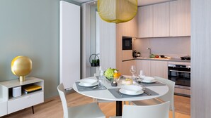 A modern dining area with a round table set for four, a kitchen in the background, and a unique yellow pendant light. - The Central City - Luxury ApartHotel (Luxembourg City)