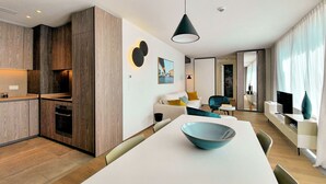 A modern kitchen with wooden cabinets, a dining area with a white table and blue bowl, and a living area with a sofa and a teal armchair. - The Central City - Luxury ApartHotel (Luxembourg City)