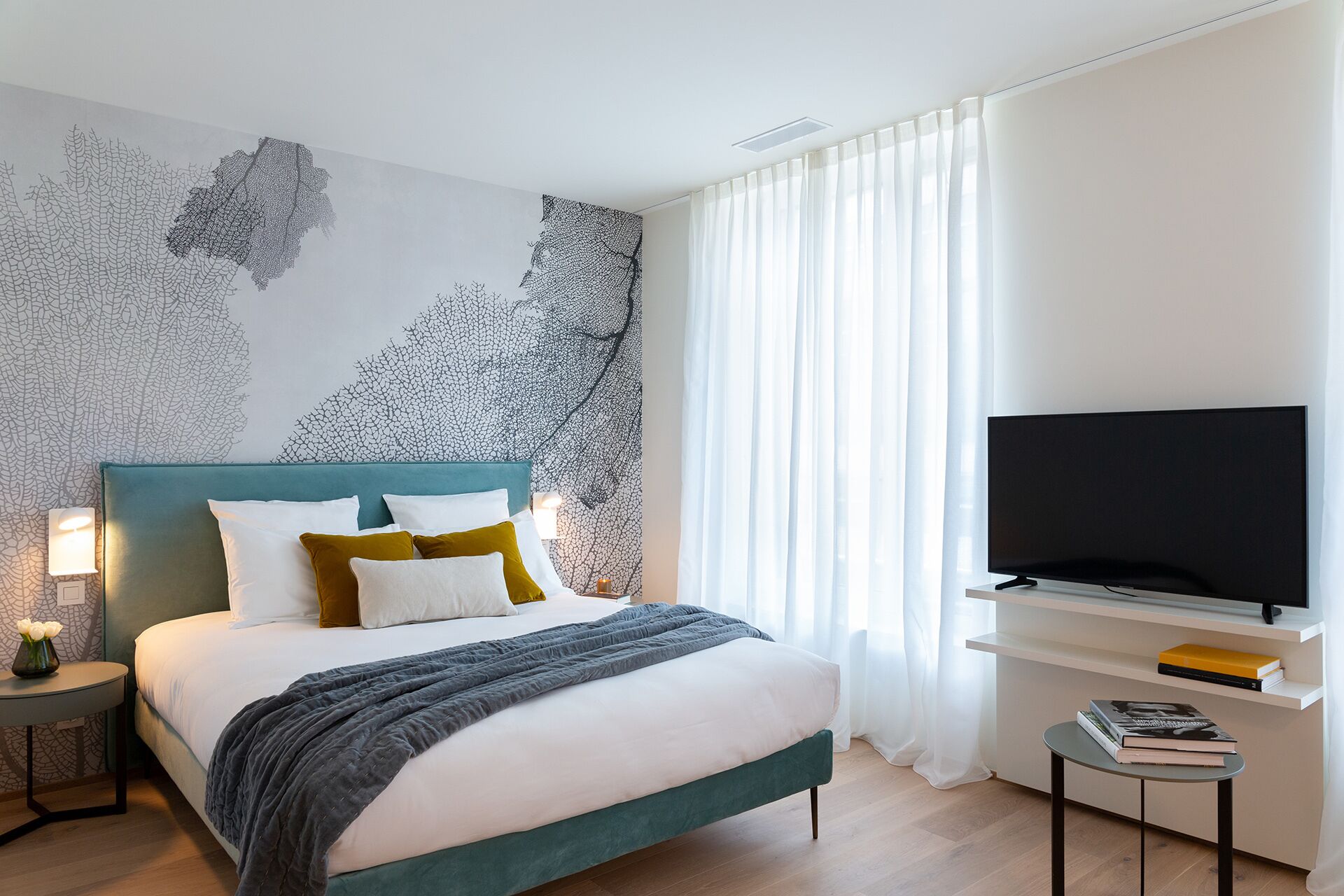 Une chambre d’hôtel moderne équipée d’un lit, d’un téléviseur à écran plat, d’une petite table avec des livres et d’un mur orné d’un motif représentant des feuilles.