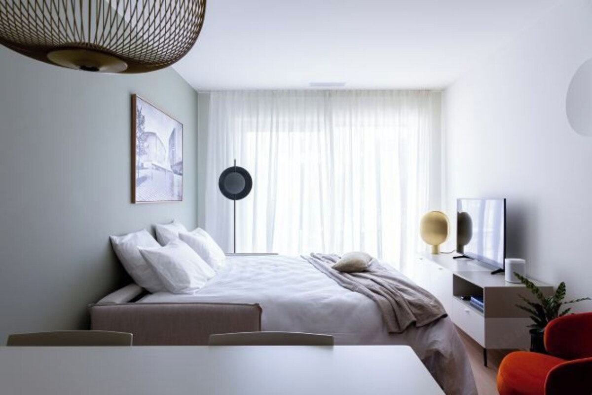 A modern hotel room with a bed, bedside table, TV, and a dining area with chairs.
