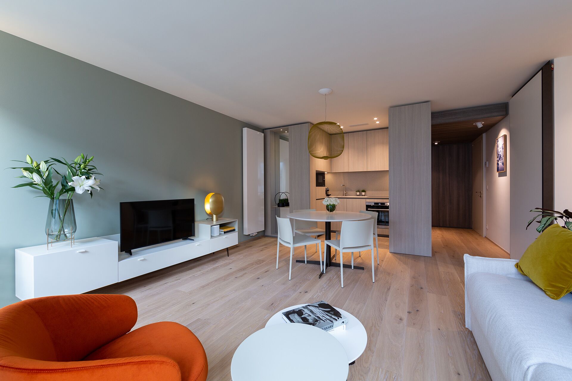 Uma sala de estar moderna com televisão de tela plana, poltronas laranja, um sofá branco e um espaço de jantar com uma mesa redonda.