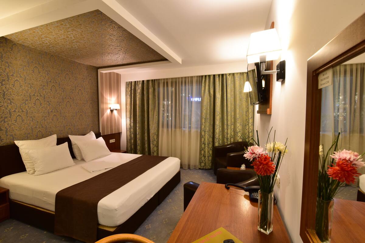 A hotel room with a large bed, a desk with a chair, a desk lamp, a vase with flowers, and a mirror.
