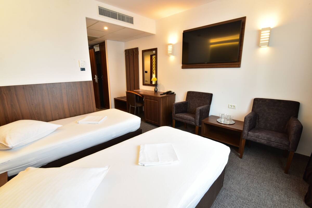 A hotel room with two single beds, a desk, two armchairs, a TV, and a mirror.
