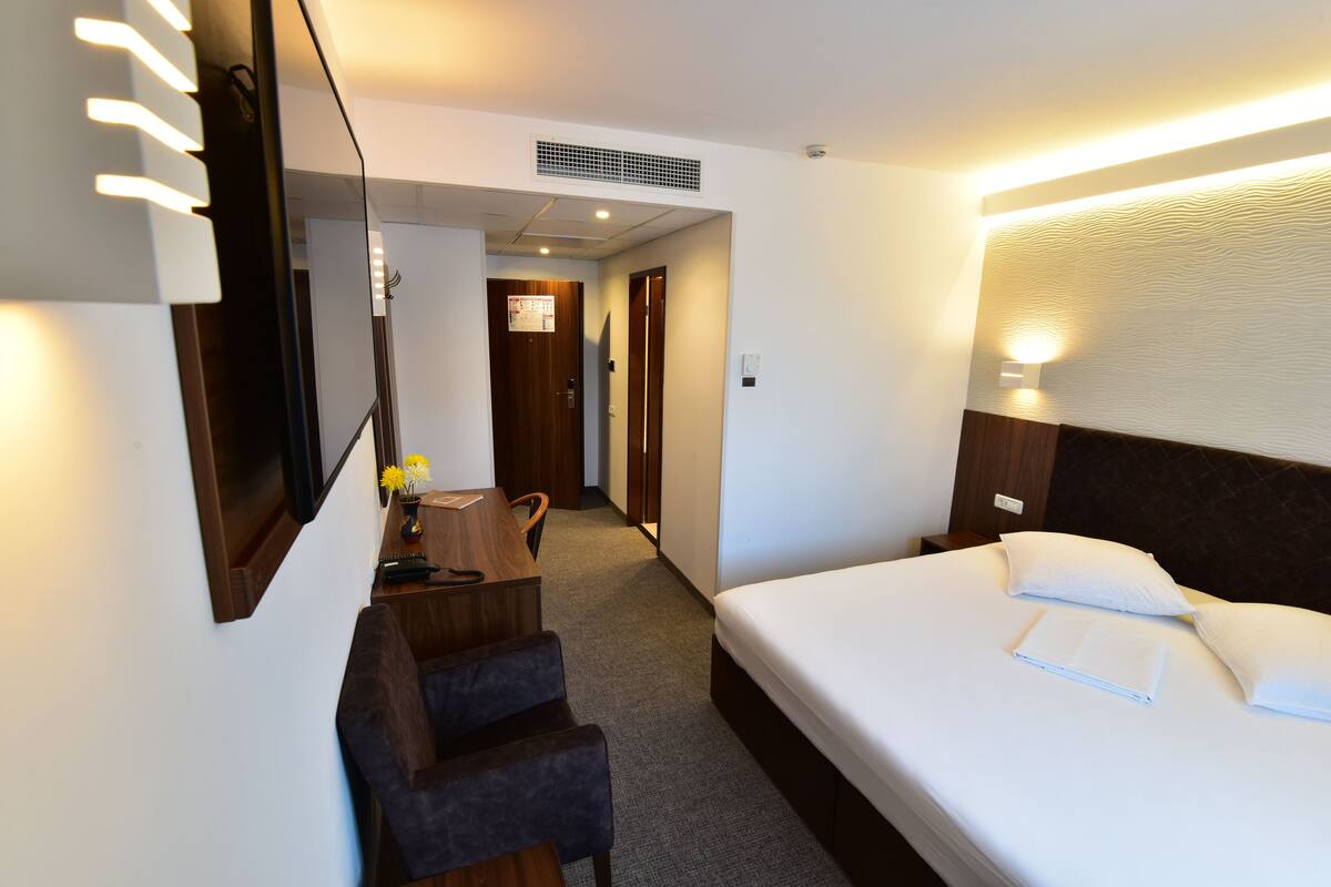 A hotel room with a bed, a desk with a chair, a mirror, and a nightstand.