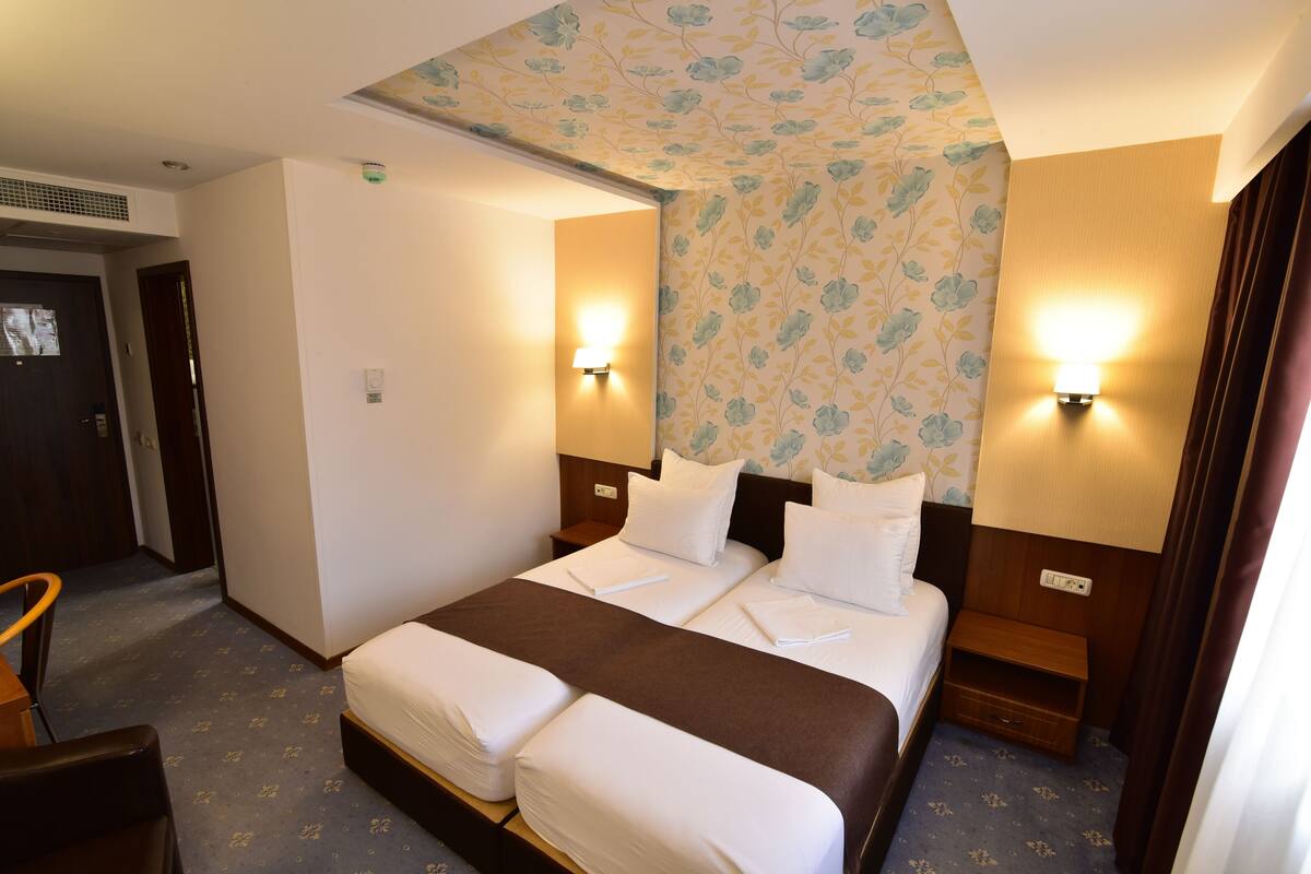 A hotel room with a double bed, two bedside tables with lamps, a wooden nightstand, a headboard with floral wallpaper, and a carpeted floor.