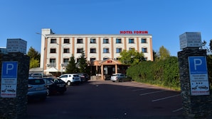 Hotel Forum, un hotel con entrada central, varios espacios de estacionamiento y un cielo azul despejado.