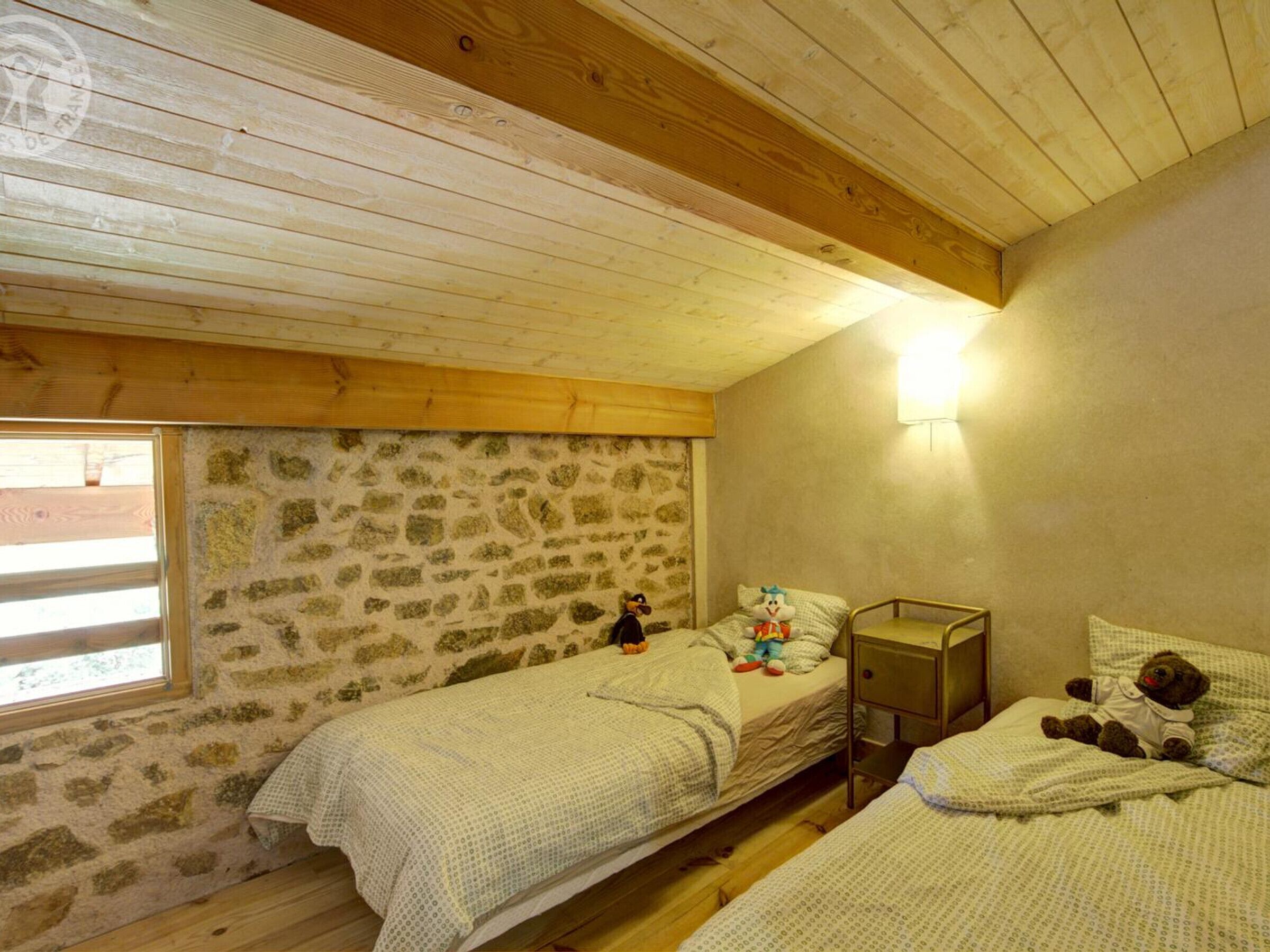 Una habitación acogedora con dos camas individuales, una pared de piedra, vigas de madera, una ventana y una mesita de noche con lámpara.