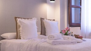 Un lit bien fait, avec du linge de lit blanc, deux oreillers et une serviette pliée. Une lampe est fixée au mur au-dessus du lit, et une fenêtre avec des rideaux se trouve à droite.