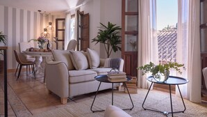 Un salon comprenant un canapé, une table basse et un coin repas. On y trouve des plantes, un lustre et une vue sur la ville.