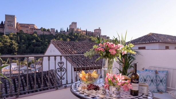 Un balcon avec une table décorée de fleurs, de fruits et d’une bouteille de vin, donnant sur une ville historique aux bâtiments qui rappellent des châteaux.