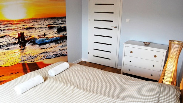 A bedroom with a bed, a white dresser, and a wall mural of a sunset over the ocean. - HelApartamenty - Apartament Bałtyk (Hel)