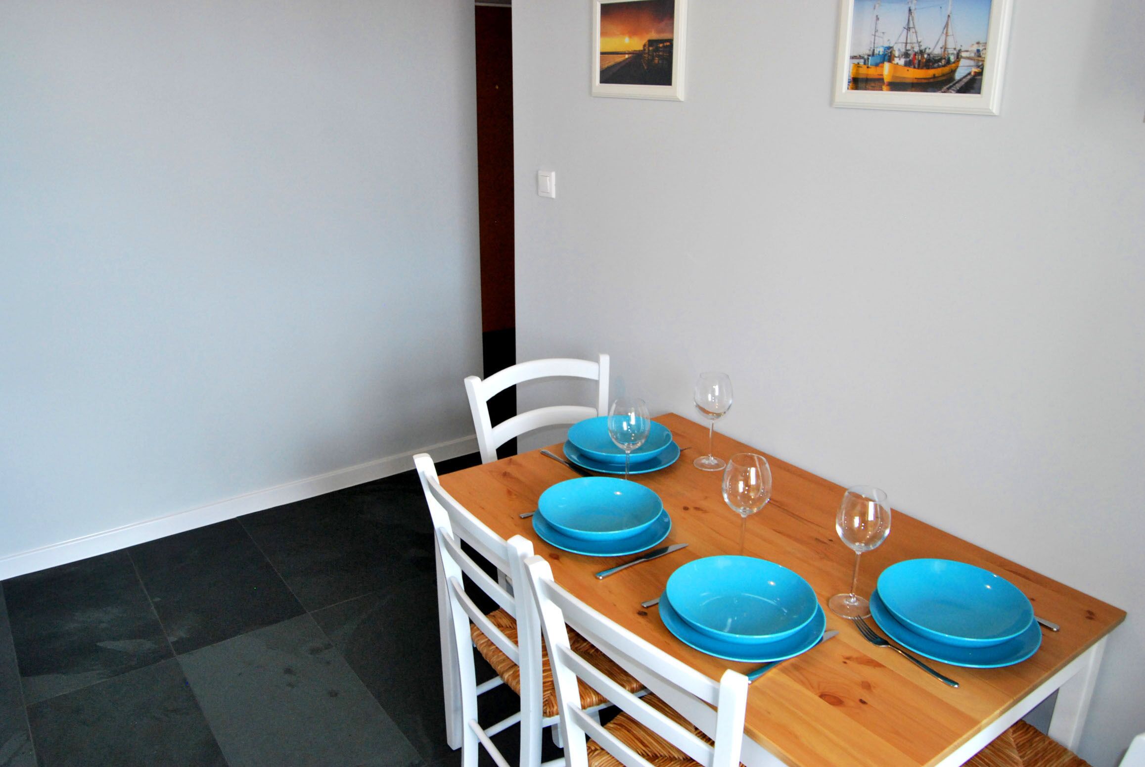 A dining table set with blue plates, wine glasses, and white chairs in a room with a tiled floor and two framed pictures on the wall.