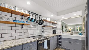 A modern kitchen with grey cabinets, a granite countertop, and a built-in oven. There are shelves above the counter holding dishes and cookware. - Island Oasis l Steps to the Ocean l 2 Pools (St. Augustine)