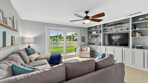 A living room with a grey sofa, a ceiling fan, a TV, and a bookshelf. - Island Oasis l Steps to the Ocean l 2 Pools (St. Augustine)
