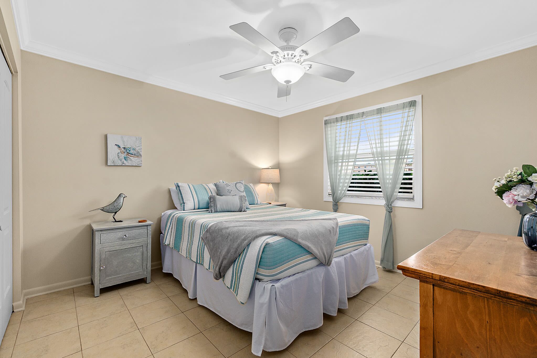 A bedroom with a bed, nightstand, ceiling fan, window with curtains, and a vase with flowers.