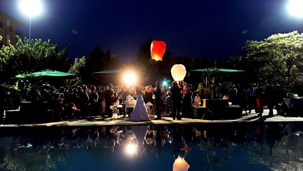 A poolside event with a crowd, a reflective pool, and bright lights in the background.