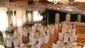 A banquet hall with round tables covered in white tablecloths and chairs, a checkered floor, and decorative plants.