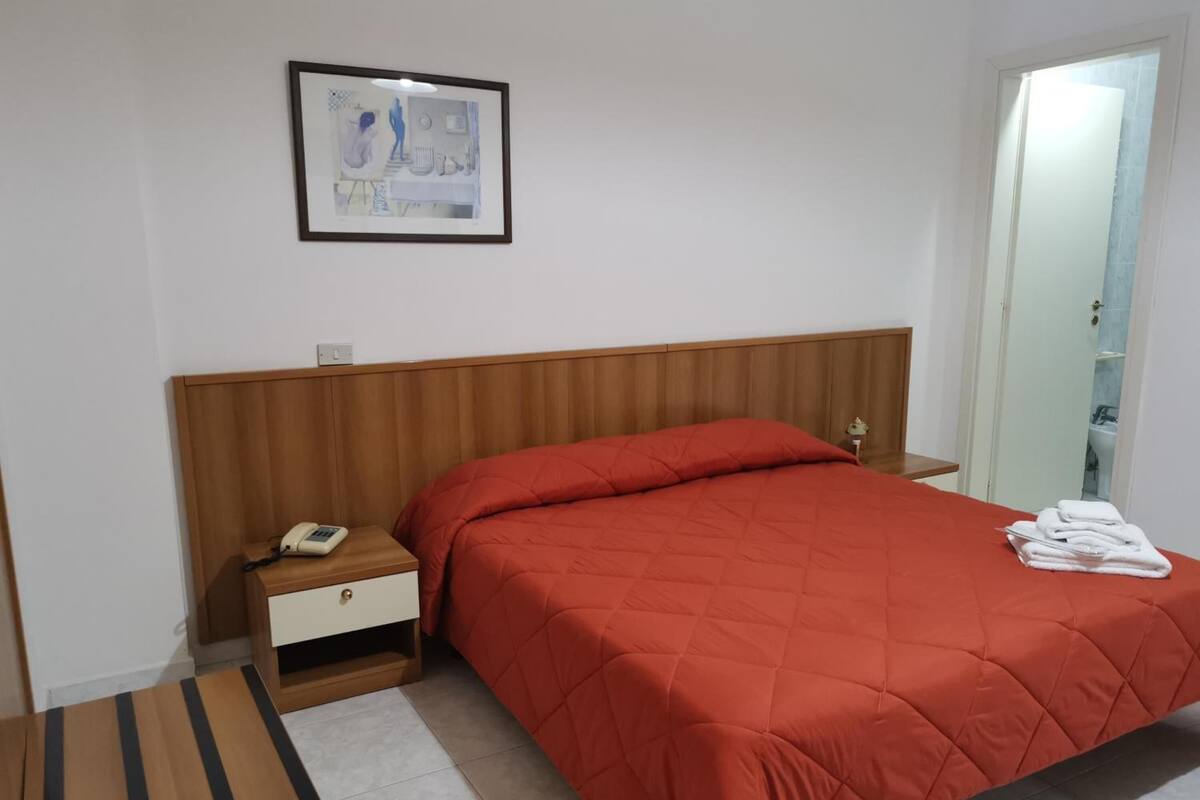 Siististi järjestetty hotellihuone, jossa on puinen sängynpääty, yksihenkinen sänky punaisella peitteellä, yöpöytä, jossa on puhelin, ja seinällä oleva kehystetty kuva.