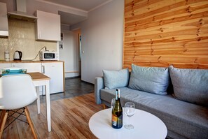 A modern living room with a grey sofa, a wooden wall, a white table with a bottle and glass, and a kitchen area with a microwave and kettle. - HelApartamenty - Apartament Baltyk II (Hel)