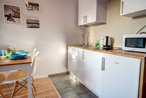 A compact kitchen with white cabinets, a microwave, and a sink. There are framed pictures on the wall and a dining table with chairs. - HelApartamenty - Apartament Baltyk II (Hel)