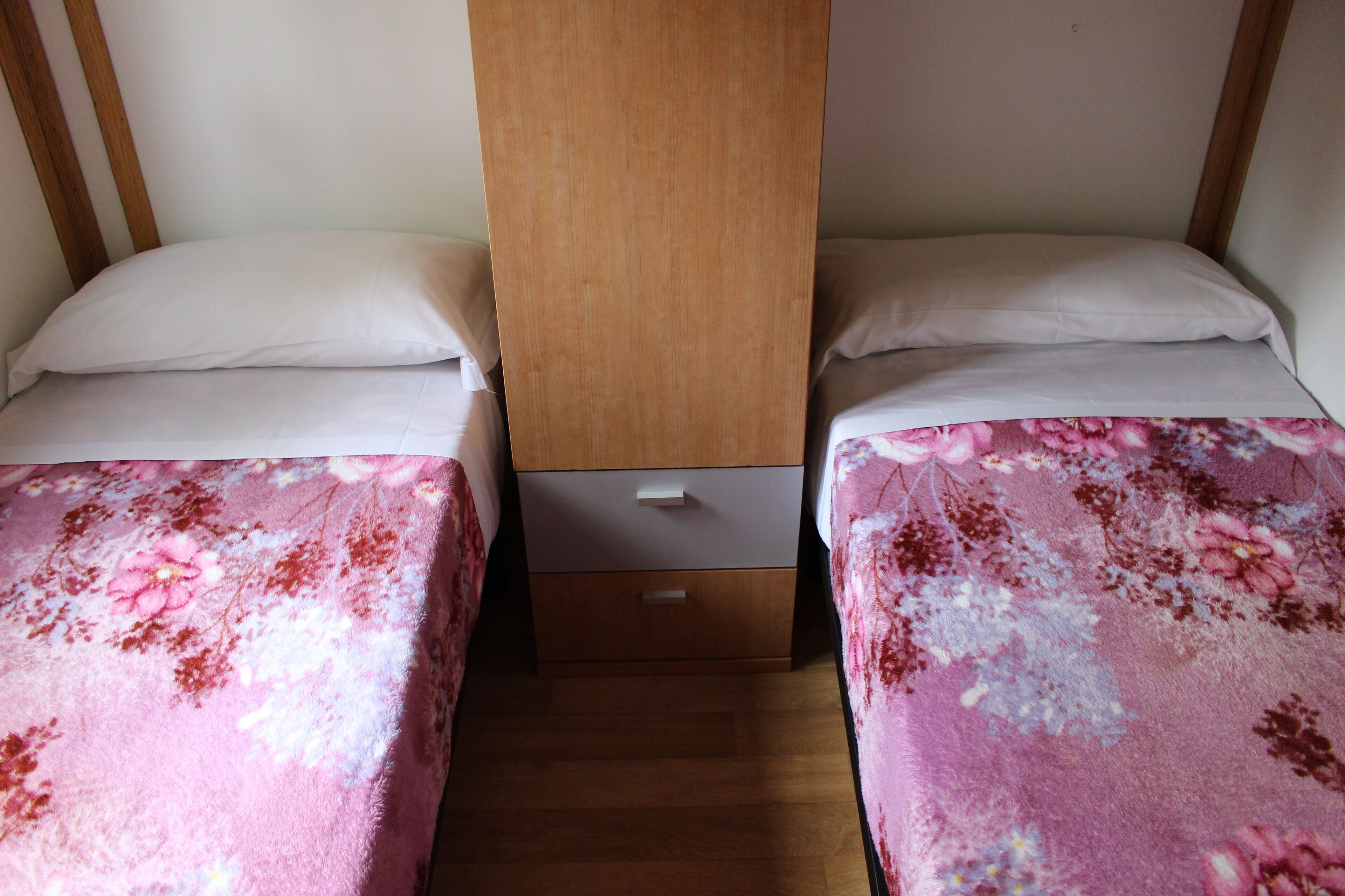 A bedroom with two beds, each with a floral bedspread, a wooden wardrobe, and a small drawer unit.
