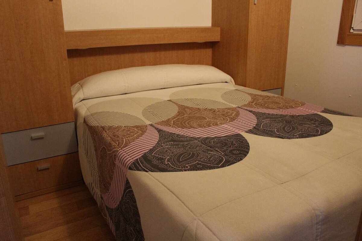 A bed with a patterned bedspread, a wooden headboard, and a small drawer unit.